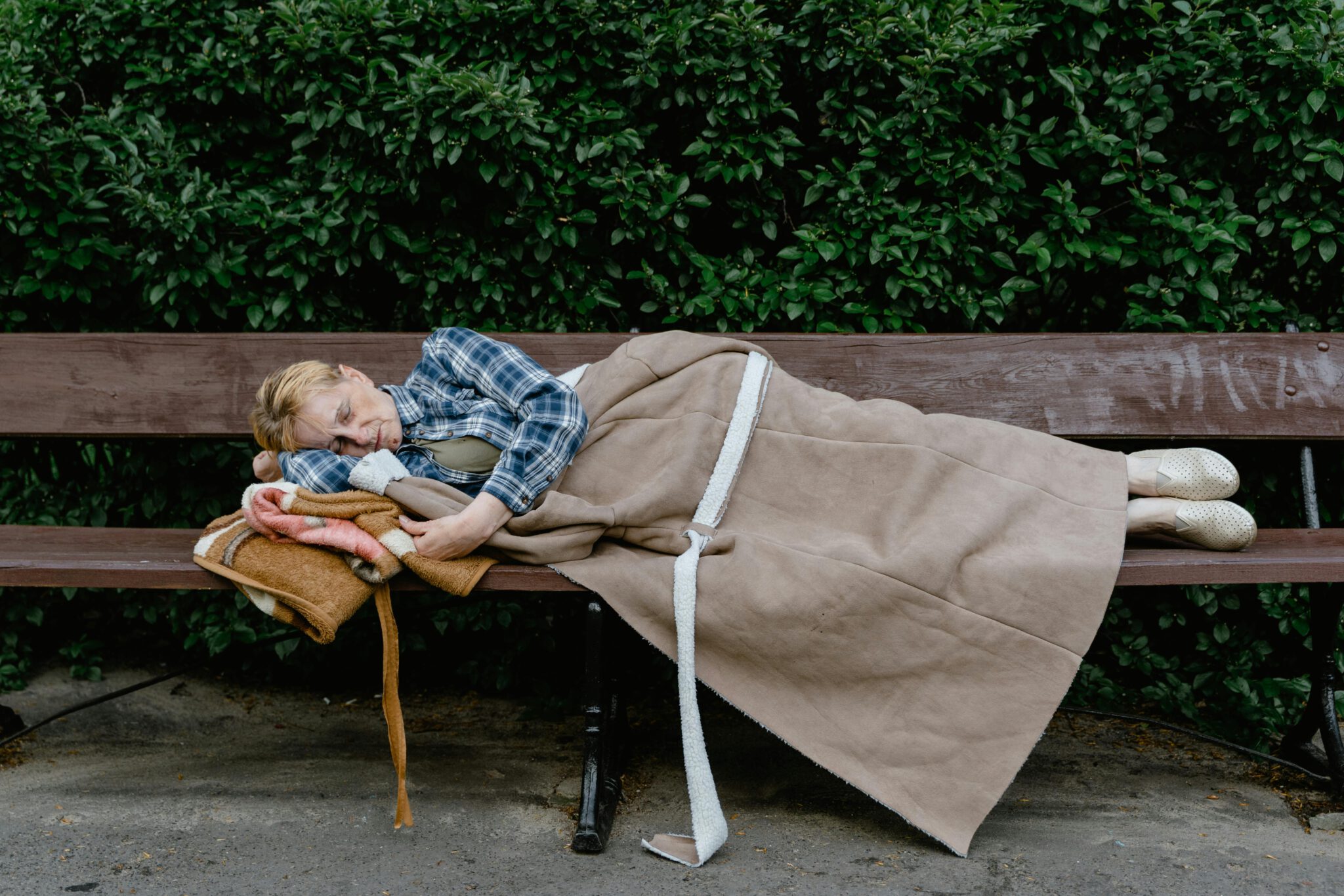 An elderly woman wrapped in a blanket sleeping on a wooden bench outdoors, surrounded by greenery.