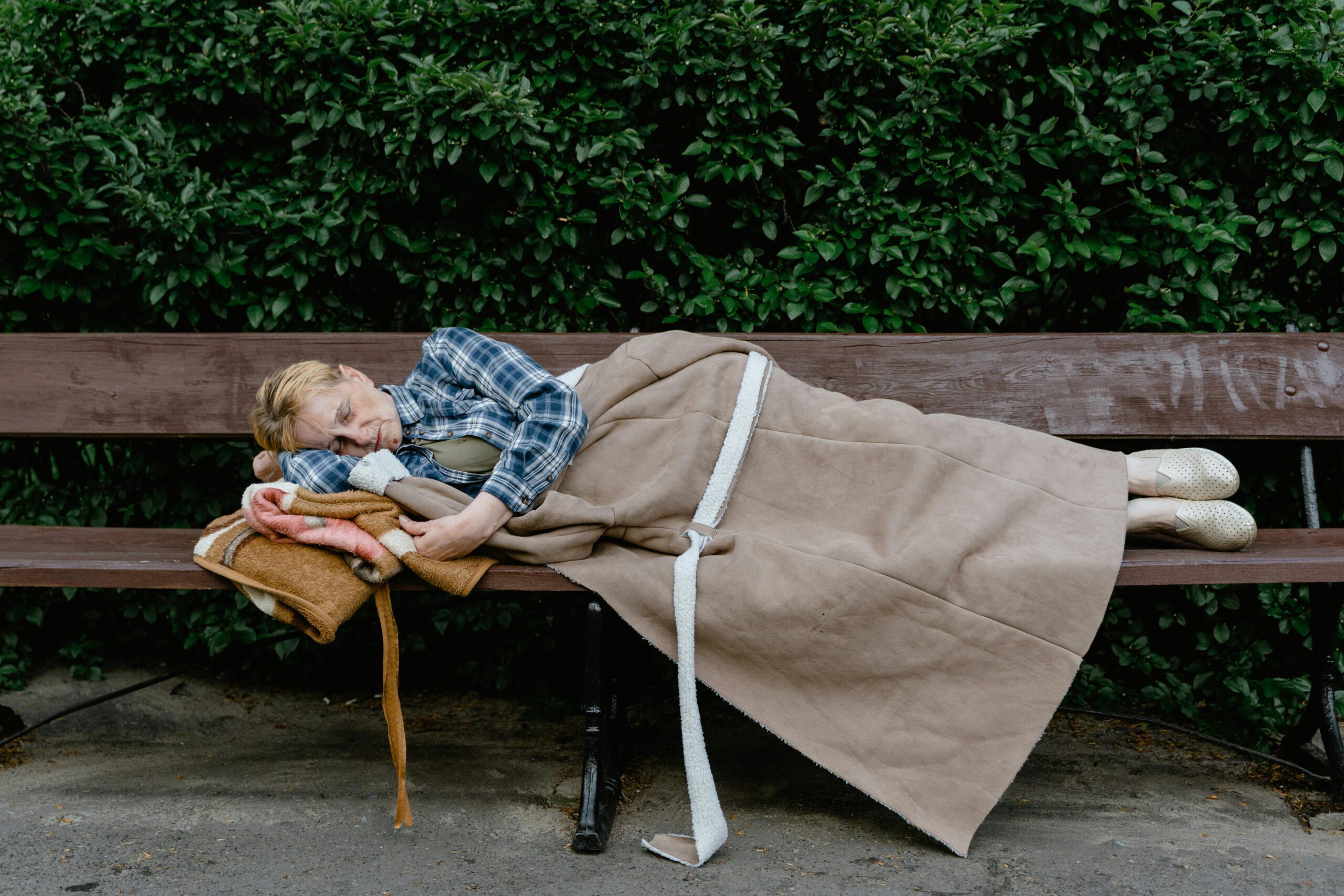 An elderly woman wrapped in a blanket sleeping on a wooden bench outdoors, surrounded by greenery.